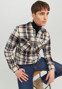 Chemise à manches longues à carreaux dans des tons crème, marine et marron ; dotée de deux poches poitrine, d'un boutonnière à l'avant et d'un col. Associée à un jean sombre.