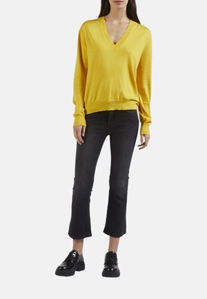 Donna che indossa un maglione giallo con scollo a V, jeans neri corti e scarpe nere con lacci, in piedi davanti a uno sfondo semplice.