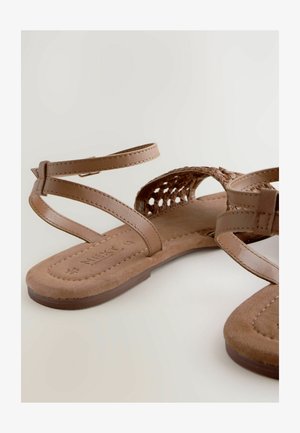 Brune sandaler med fletdesign, justerbare ankelremme og en flad, polstret sål. Materialet ser ud til at være syntetisk med en blød tekstur.