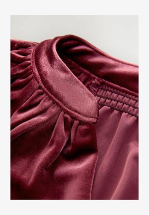 Burgunderrött satinmaterial med en veckad krage och detalj i elastisk smock. Mjuk, slät textur med lätt glans och rena kanter.