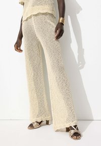 Ensemble tricot beige composé d'un haut ample et d'un pantalon large à bords festonnés. Le modèle porte des sandales à talons avec des lanières.