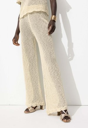 Ensemble tricot beige composé d'un haut ample et d'un pantalon large à bords festonnés. Le modèle porte des sandales à talons avec des lanières.