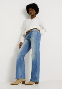 Jeans de mezclilla de color azul claro con un diseño acampanado, combinados con una blusa blanca de manga larga con detalles de encaje y un cinturón marrón.