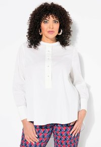 Chemise blanche à manches longues avec un col montant et un plastron avant agrémenté de boutons noirs. Portée avec un pantalon multicolore à motifs.