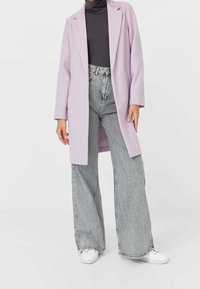 Lila lång kappa med urklippta slag, bärs över en svart polotröja, parat med högmidjade vida grå jeans och vita sneakers.