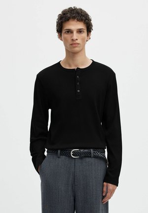 Jeune homme aux cheveux bouclés portant un shirt henley noir à manches longues et un pantalon gris texturé avec une ceinture tressée, debout devant un fond uni.