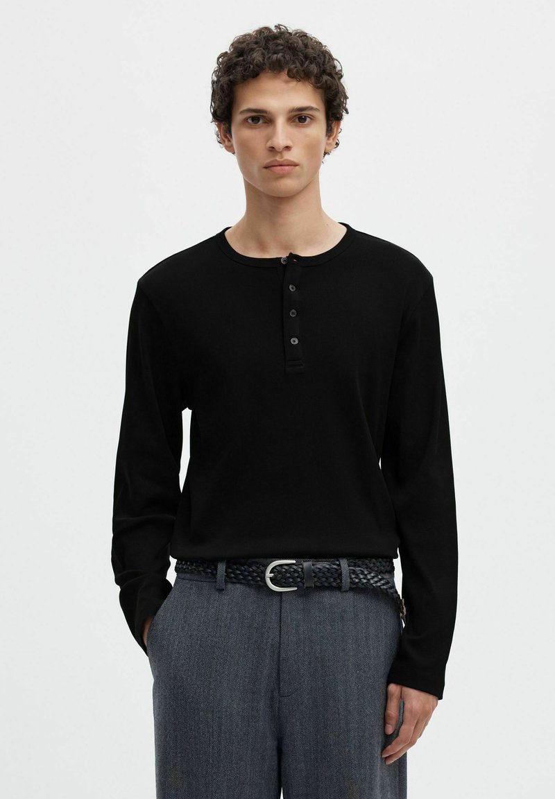 Jeune homme aux cheveux bouclés portant un shirt henley noir à manches longues et un pantalon gris texturé avec une ceinture tressée, debout devant un fond uni.