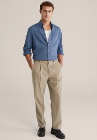 Kék, gombos ing mellzsebbel, világosbézs pliszírozott nadrággal és fekete cipővel párosítva. Simább anyag, testre szabott szabás.