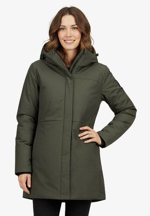 Vrouw met golvend bruin haar die een lange olijfgroene winterjas met capuchon draagt, glimlachend en poserend met één hand op haar heup.