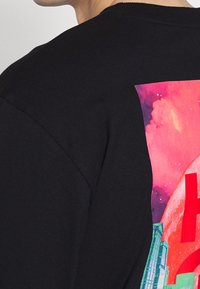 Schwarzes Shirt mit buntem Grafikdesign, das einen roten Planeten, einen rosa Himmel und eine grüne Stadtlandschaft auf dem oberen Rücken einer Person zeigt.