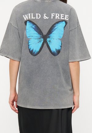 Personne portant un T-shirt gris délavé avec un grand papillon bleu et le texte "WILD & FREE" dans le dos.