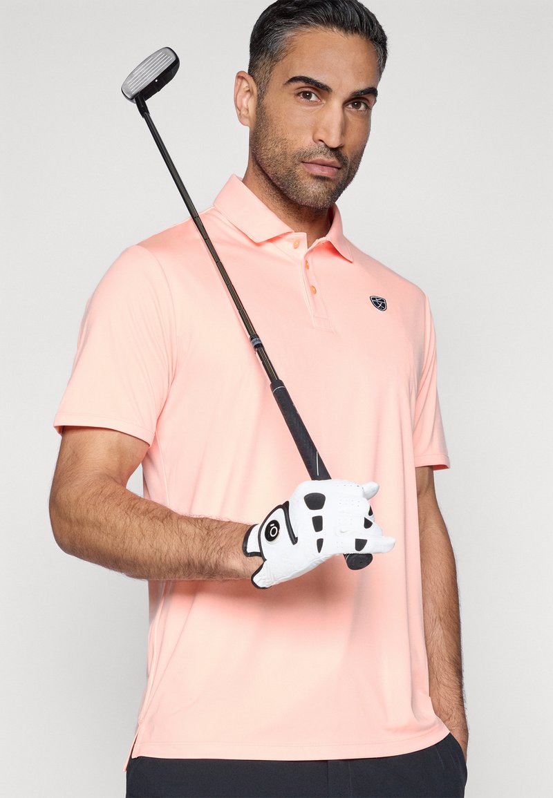 Polo rose avec col et manches courtes. Le modèle tient un club de golf, portant un gant blanc avec des accents noirs. Design simple.