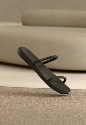 Sandale negre Crocs cu două curele minimaliste plutind deasupra unui covor bej într-o cameră cu tonuri neutre și mobilier moale în fundal.