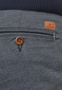 Gros plan sur la poche d'un pantalon en tissu gris texturé, avec un bouton marron et une étiquette de marque en cuir Jack & Jones.