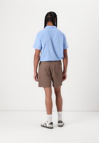 Chemise légère à manches courtes de couleur bleu clair associée à un short marron avec taille élastique. Portée avec des chaussettes blanches et des sandales à rayures noires.