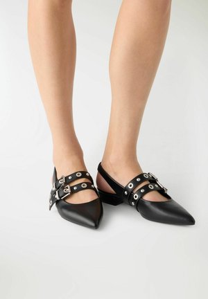Gambe con scarpe basse nere con punta appuntita, doppia fibbia e dettagli in occhiello argentati su sfondo chiaro uniforme.