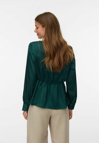 Bluza od satena u tirkiznoj boji s dugim rukavima, suženim strukom i ležernim krojem. Glatka tekstura i diskretne nabore na leđima, u kombinaciji s bež hlačama.