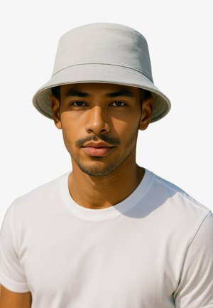 Młody mężczyzna z jasnym zarostem, noszący jasnoszary kapelusz typu bucket i jednolitą białą koszulkę, zwrócony przodem z neutralnym wyrazem twarzy.