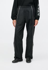 Άτομο που φοράει μαύρο παντελόνι φόρμας με λευκές ρίγες, μαύρα αθλητικά παπούτσια και μαύρο μακρυμάνικο μπλουζάκι με λευκό κείμενο στο μανίκι.