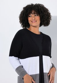 Frau mit lockigem Haar, die einen schwarz-weißen und grauen Blockstreifen-Cardigan über einem schwarzen Oberteil trägt, lächelt und schaut zur Seite.