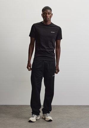 Sort mand står mod en ensfarvet lys væg iført sort t-shirt og joggingbukser med hvide sneakers på gråt gulv.