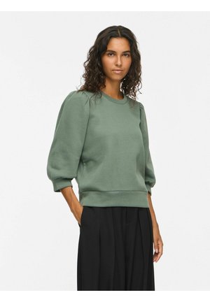 Femme aux cheveux bouclés portant un pull vert à manches bouffantes et un pantalon plissé noir, debout avec une main dans la poche sur fond blanc.