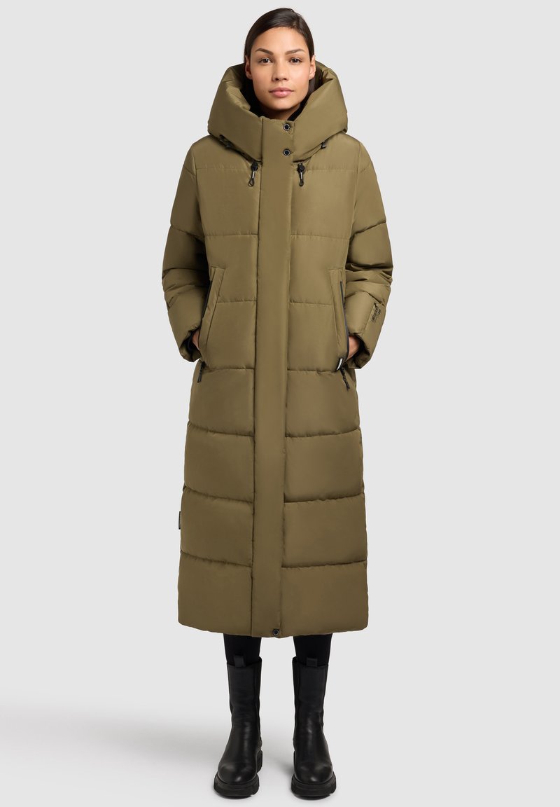 Olivgrön lång pufferjacka med en stor huva, fickor med dragkedja och ett kviltat mönster. Har knappar fram och en slät textur.