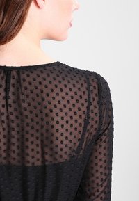 Černý průsvitný top s texturou puntíků, se zaobleným výstřihem a zapínáním na knoflíky. Hladký materiál s jemnými matnými akcenty.