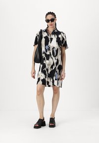 Rochie cu mâneci scurte și nasturi, cu un model abstract în culorile negru, alb și albastru, prezentând o croială dreaptă și purtată cu o geantă neagră și sandale masive.