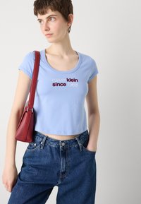 Light blue cotton cropped t-shirt with a round neckline and short sleeves, featuring dark red text “calvin klein since 1968.” Paired with high-waisted denim jeans and a red handbag.
