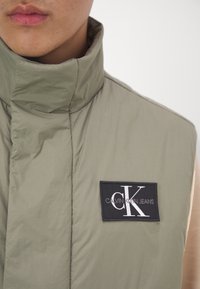 Světle olivově zelená vesta s vysokým límcem, na levé straně s černou nášivkou s logem "Calvin Klein Jeans" a bílým nápisem.