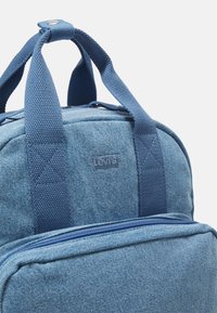 Mochila de denim en azul medio con tejido texturizado, asas de cinta resistentes, un bolsillo frontal con cremallera y un sutil detalle de logo.