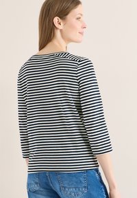 Femme aux cheveux bruns lisses portant un haut noir et blanc à rayures à manches longues et un jean en denim bleu, regardant par-dessus son épaule contre un mur uni.