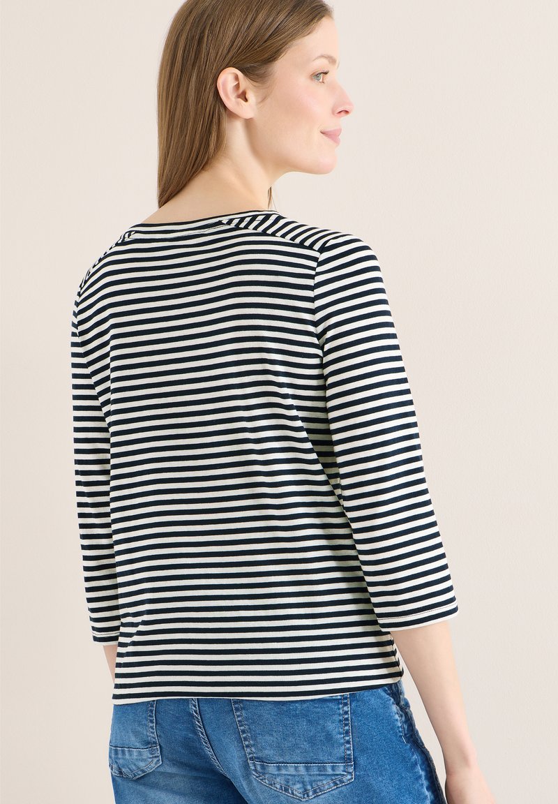 Femme aux cheveux bruns lisses portant un haut noir et blanc à rayures à manches longues et un jean en denim bleu, regardant par-dessus son épaule contre un mur uni.