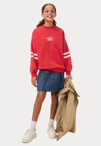 Felpa rossa con strisce bianche, gonna di jeans e sneakers bianche. La felpa presenta una stampa grafica e polsini a coste. Giacca beige in mano.
