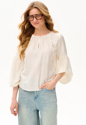 Kobieta z długimi falującymi włosami, nosząca dużych rozmiarów okulary przeciwsłoneczne w kolorze żółwia, kremową bluzkę z falbankami i jasnoniebieskie dżinsy, ręka w kieszeni.