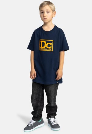 Marineblauwe katoenen T-shirt met gele DC-logo, gecombineerd met donkere spijkerbroek en grijze sneakers met turquoise accenten.