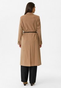 Beige lange jas met een verstelbare taille, getailleerde schouders en zijsplitten. Draagt over een brede donkerblauwe spijkerbroek en bruine schoenen. Glad textuur.