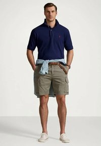 Ναυτικό πόλο μπλουζάκι, καφέ κοντομάνικες παντελόνες cargo με τσέπες, ανοιχτό μπλε πουκάμισο δεμένο στη μέση, και λευκά αθλητικά παπούτσια. Απλός, casual σχεδιασμός.