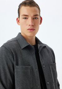Camisa de tejido gris con cuello tipo solapa, botones a presión y dos bolsillos frontales. Material texturizado con un estampado sutil.