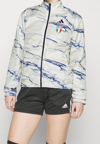 Adidas Italia-jacka i ljuskräm med blå marmorering, dragkedja fram, hög krage, elastiska manschetter, matchad med svarta shorts med logotyp.