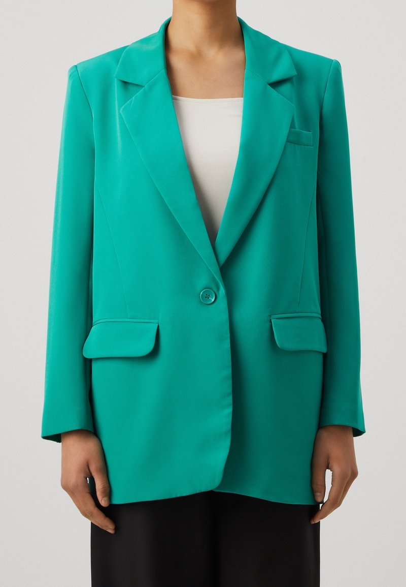 Blazer turquesa con un diseño entallado, solapas de muesca, dos bolsillos frontales y cierre de un solo botón. Textura de tela suave.