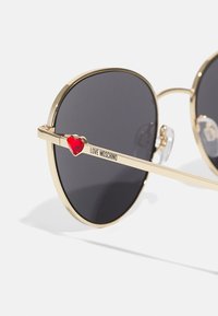 Gold-framed sunglasses with dark lenses, featuring a prominent red heart accent on the arm and the text "LOVE MOSCHINO."