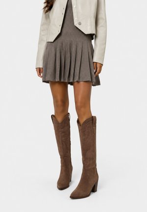 Kvinde iført en ribbet plisseret brun nederdel, beige kort jakke og knæhøje brune ruskindsstøvler med hæl på hvid baggrund.