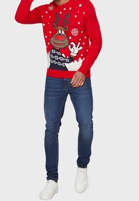 Pull en tricot rouge avec un motif de renne, des flocons de neige blancs et un ourlet côtelé. Associé à un jean bleu foncé et des sneakers blancs.