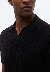 Μαύρο πλεκτό πόλο πουκάμισο με υφασμένο μοτίβο καλωδίου, κοντά μανίκια και στάνταρ γιακά με άνοιγμα σε σχήμα V.