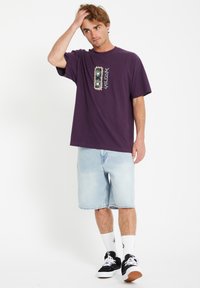 Paarse katoenen T-shirt met een grafische print, gecombineerd met lichtblauwe denim shorts. Model draagt zwarte sneakers en witte sokken.