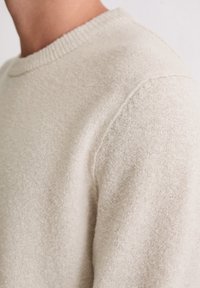 Jasno-beżowy sweter z okrągłym dekoltem, wykonany z miękkiego, teksturowanego materiału. Posiada ryflowany dekolt z delikatnymi detalami przeszycia w okolicy ramion.