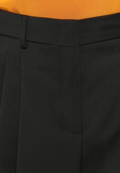 Pantaloni neri su misura con fronte piatto, texture in tessuto intrecciato, leggeri pizzicotti e vita standard. Lo sfondo presenta un tessuto arancione.