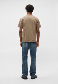 Barna pamut póló rövid ujjakkal, elnyűtt kék farmernadrággal párosítva. A magas sarkú cipők feketék, a háttér világos.